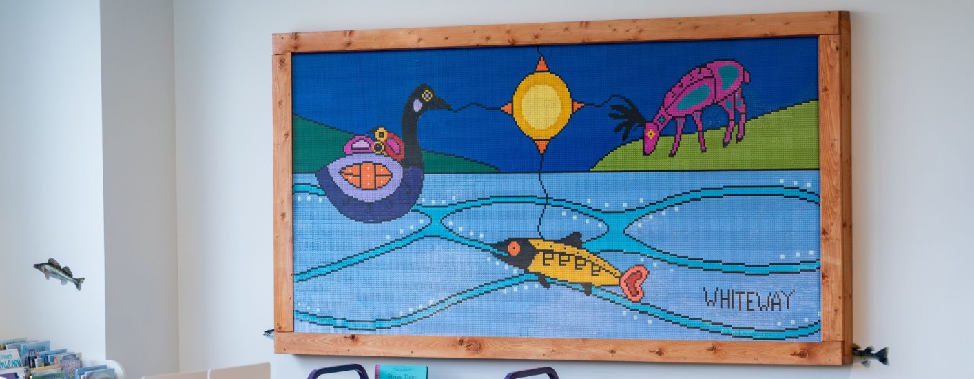 Large framed mosaic-style artwork mounted on a library wall, depicting a stylized landscape with a loon, fish, grazing animal, and bright sun connected by winding lines across blue water. The artwork includes the word “WHITEWAY” in the lower right corner 