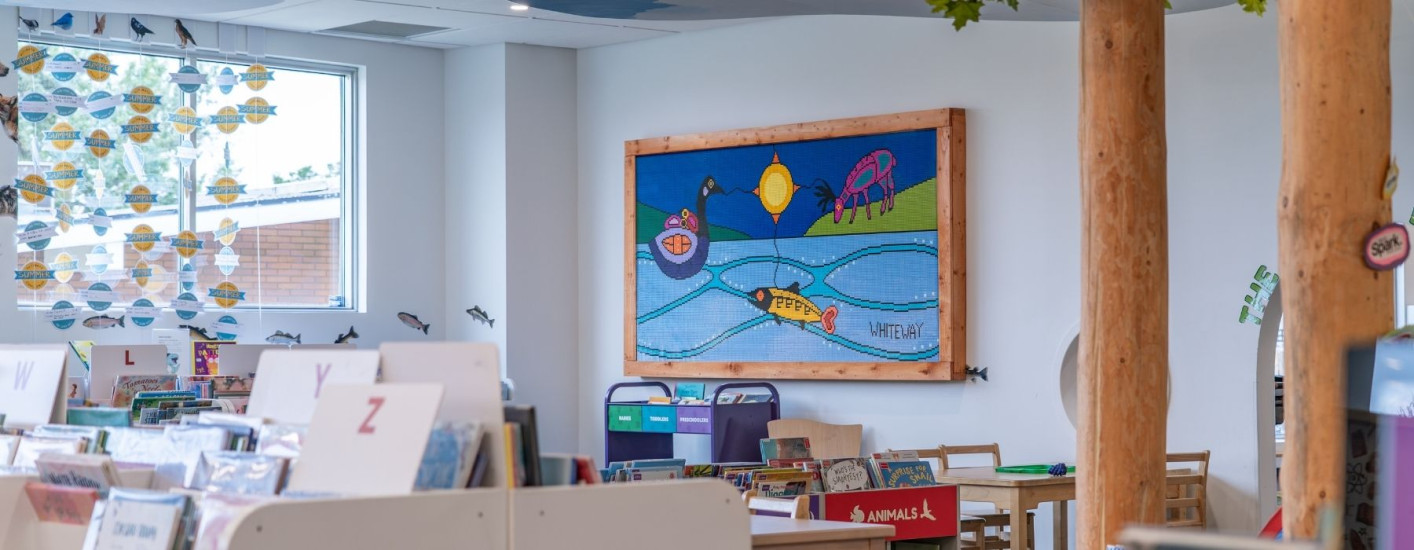 Children's library area with low bookshelves, alphabet dividers, and small reading tables. A large framed colourful artwork depicting fish, birds, and a landscape hangs on the wall.