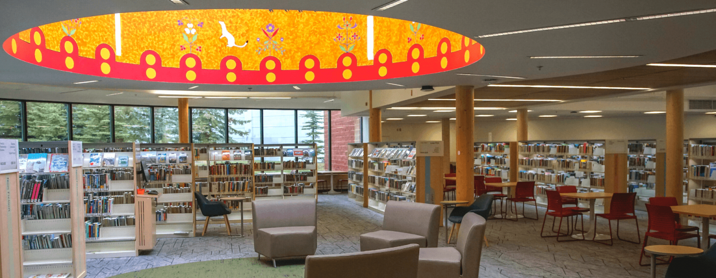 A spacious Library interior with bookshelves, seating areas, and tables. A colourful circular art piece is mounted on the ceiling, adding a vibrant touch to the room.