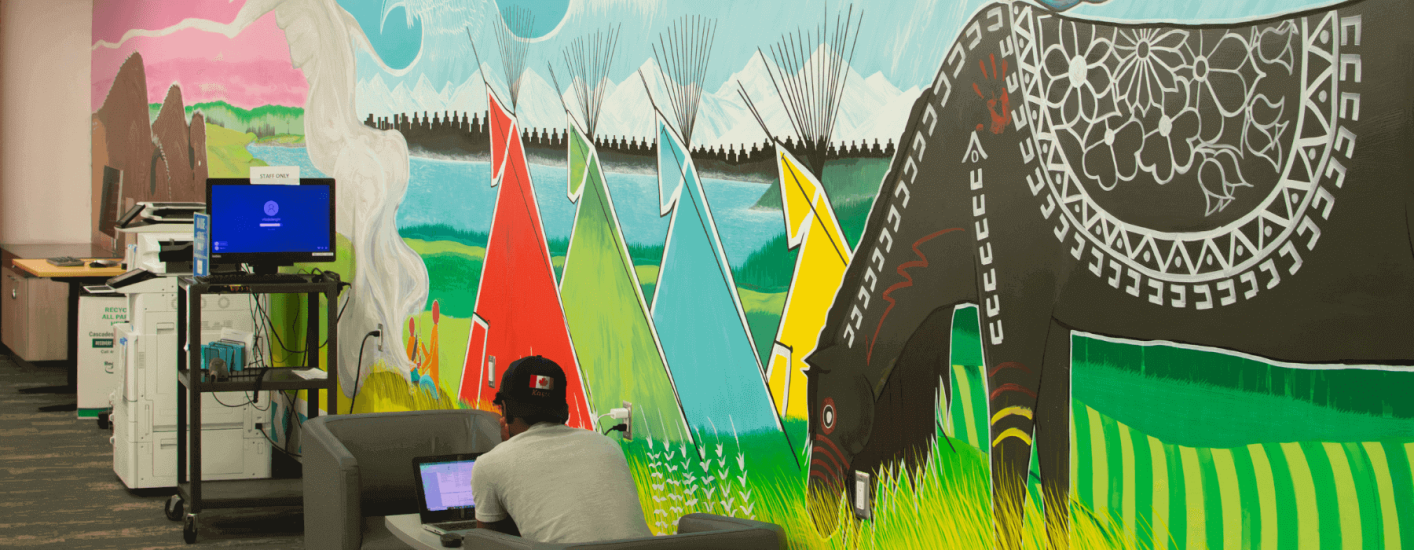 A person sits working on a laptop with their back to the camera. Behind them, a vibrant mural depicts tipis and a horse in a field. 