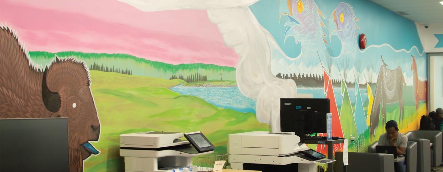 A colourful mural spans a wall, showcasing a bison, a peaceful lake scene, and Indigenous imagery. People are seated on a chair in front of the mural, near a computer station and printers.