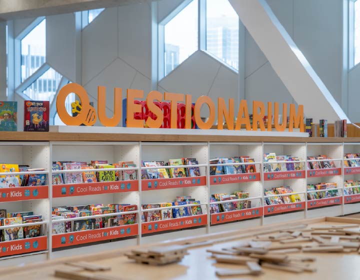 background image: Children's books displayed on library shelves beneath a large orange "Questionarium" sign.