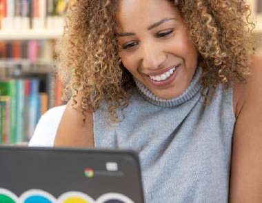 Adult using a laptop in a library with bookshelves in the background.