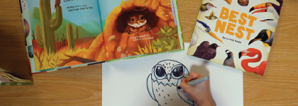 A person draws an owl while children’s books, including “Best Nest,” are open on a table.