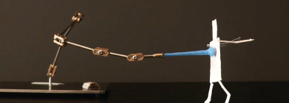 A stop-motion puppet made of wire and paper stands on a table connected to an adjustable metal arm.
