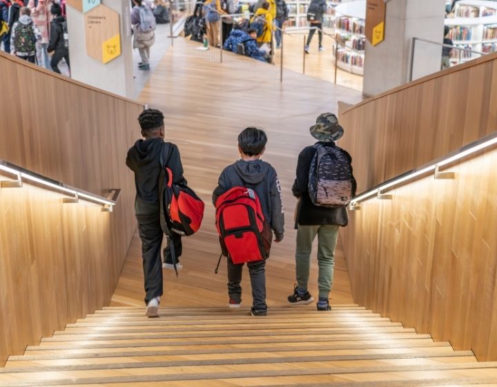 background image: Three children with backpacks walk down a wooden staircase inside a modern library.