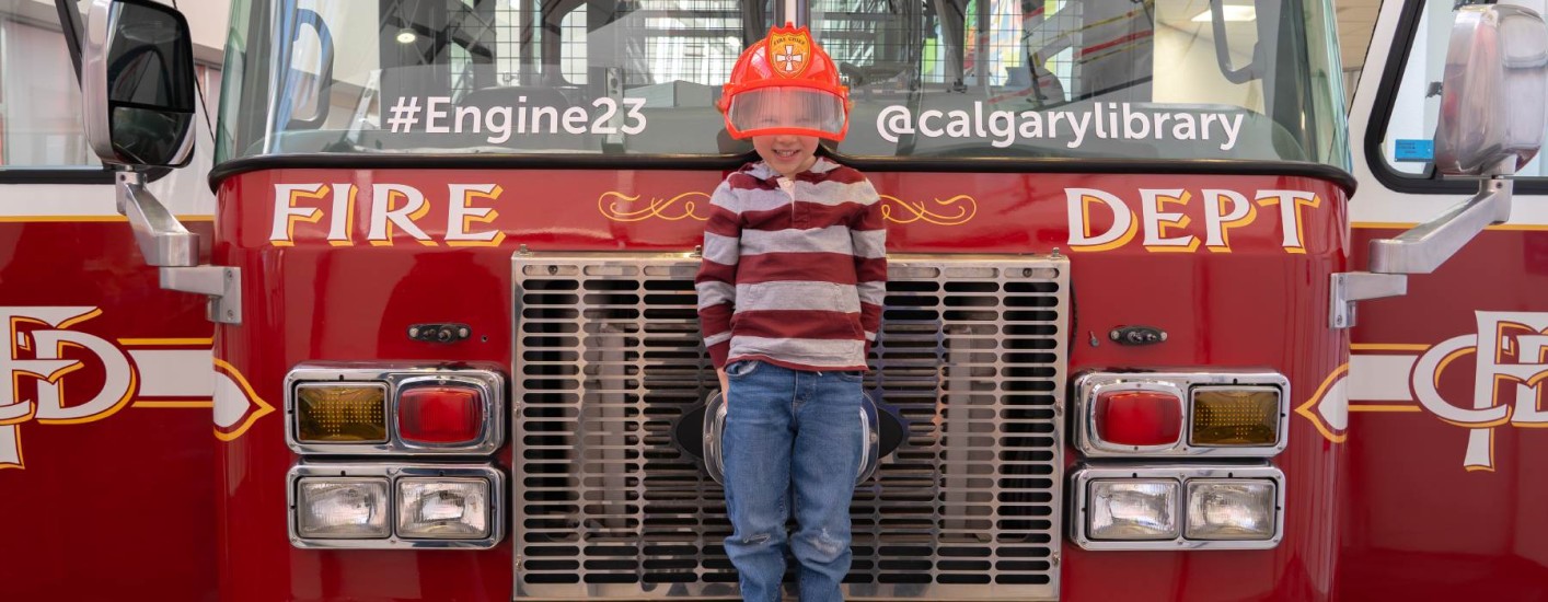 A young boy stands in front of Engine 23's firetruck. He is wearing a red firefighter's helmet and smiling at the camera. 