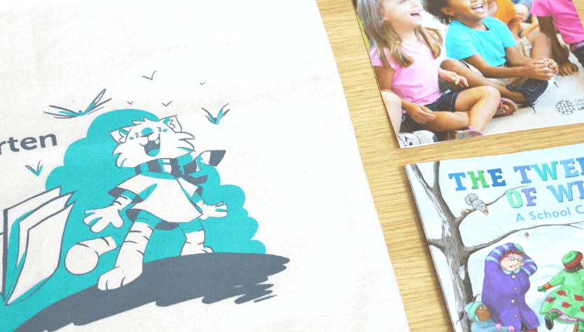 Photograph of a canvas book bag with an illustration of a cat and a book, the Calgary Public Library logo and text that reads "ready for kindergarten." There are also other reading materials on the table beside the bag. 