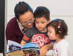 An adult reads a picture book with two young children who sit close on either side, all three focused on the pages. The scene appears warm and intimate, set against a simple indoor background.