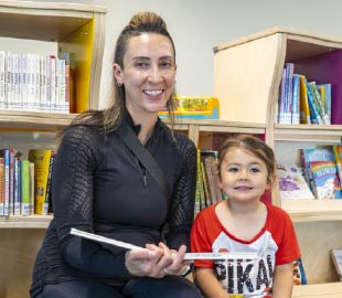 An adult and a young child sit together in a library’s children’s section, smiling at the camera. The adult holds an open book, and shelves of colourful children’s books are visible behind them.