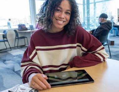 Teen seated at a table using a tablet in the library.