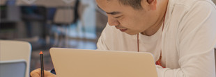 This is an image of a young man sitting at a table in Central Library. He has a laptop in front of him and writing with a pen.