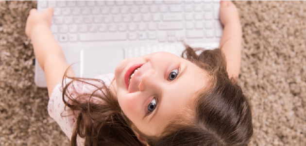 A little girl wearing a pink shirt is sitting on a carpeted floor. You are looking down at her as she smiles up at the camera. Within her hands is a laptop computer. 