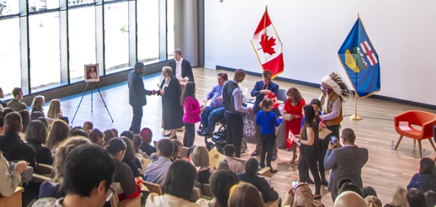 A photograph of a full auditorium. There are two flags, a Canada flag and an Alberta flag, in the background. In front of the flags, there are a group of people shaking hands. 