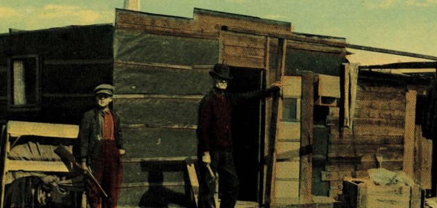 A historic photograph of a boy holding a rifle and an adult standing in front of a wooden building. 