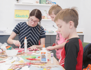 A photograph of an adult gluing someone on construction paper. She is accompanied by two boys who are also partaking in the craft.