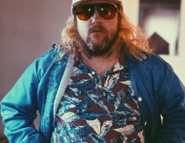 Rozsa Arts musician Carter Felker wearing sunglasses and a baseball cap posing in a blue jacket and patterned shirt.