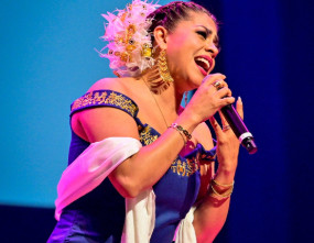 Rozsa Arts Musician Gisela Romero sings into a microphone on stage under colourful lighting.