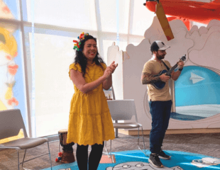 A photograph of a performer at Sage Hill Library, she is clapping and is accompanied by a male playing the ukulele behind her.