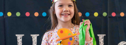 A smiling child wearing a crown, holding a paint palette and bag in front a colourful banner for a Kindergarten Book Bag Family Event