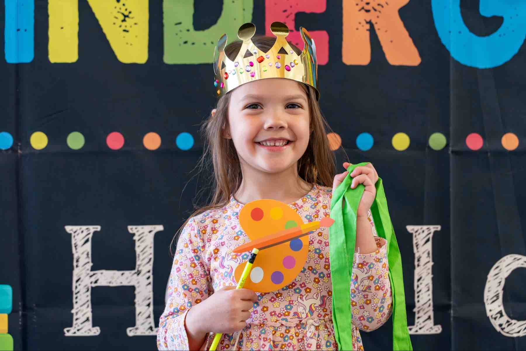 background image: A smiling child wearing a crown, holding a paint palette and bag in front a colourful banner for a Kindergarten Book Bag Family Event