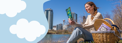 A adult sitting in front of the rive with a picnic basket of books and reading. 