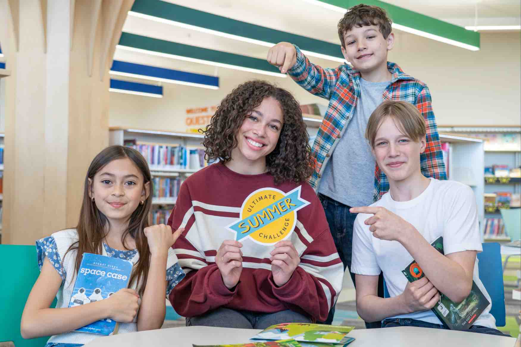 background image: Four children pose together in a Library with books and colourful displays behind them. One child holds an Ultimate Summer Challenge sign, while two others hold books and point toward the sign.