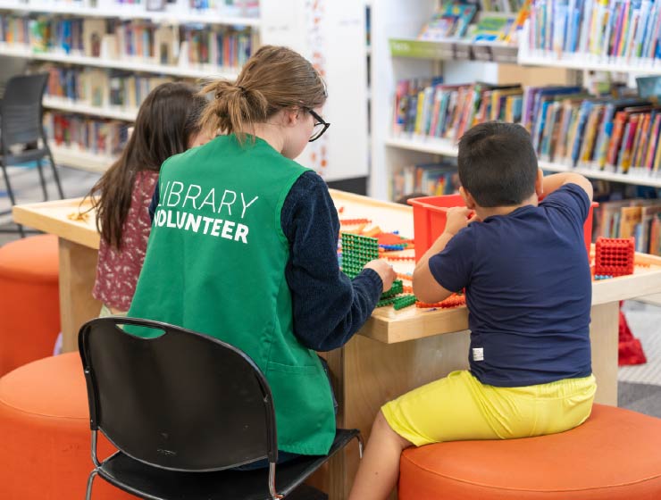 background image: Volunteer in green vest, playing at a table, building with two children as a Library "Play Champion" with bookshelves in the background