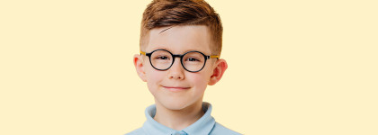 A young boy with short brown hair and round glasses, smiling slightly while wearing a light blue collared shirt, posed against a pale yellow background.