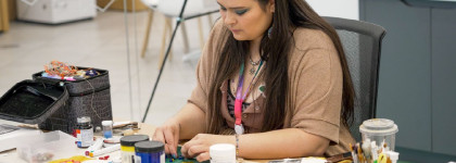 A person sits at a table covered with art supplies, carefully creating a craft project with beads, paints, and mixed-media materials.