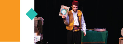 A man in a gold vest with a red hat holding up a book while on stage.