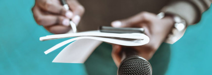Close-up of a person holding a microphone in one hand and writing on a notepad with the other, with the focus on the hands and the paper against a teal background.