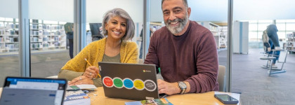 Two people working together at a table in a library study room with a laptop, books, and notebooks.