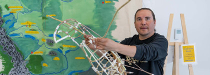 2026 Indigenous Artist in Resident with a sculpture in front of a large map for Aikahtsi Final Exhibit Opening Reception