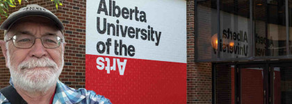 Photo of a person wearing a hat and glasses outside the Alberta University of the Arts building, with the school’s sign visible on a red and white brick wall behind them to promote Digital Alberta Art University at 100 — Some Historical Highlights event