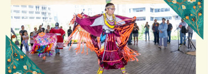 Person in colourful traditional regalia dancing at an indoor powwow while attendees watch in the background.