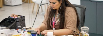 A woman is seated at a table working on beadwork, surrounded by art supplies, jars of paint, brushes, and tools. The individual is focused on their craft, with materials and colorful beads spread across the workspace.