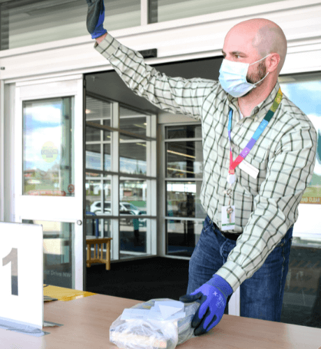 A image of a Calgary Library staff member wearing a mask and gloves and waving a someone in the distance.