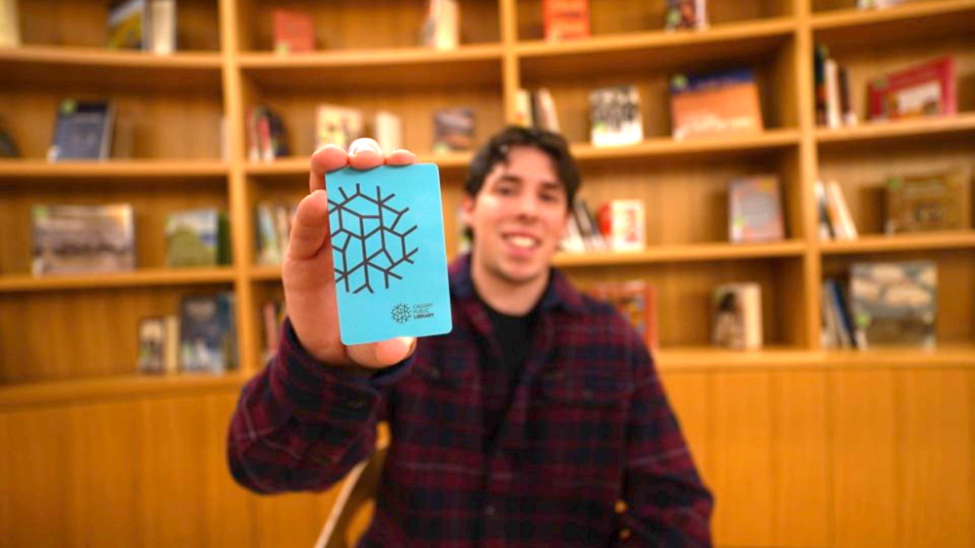 A smiling person holds up a blue Calgary Public Library card in front of bookshelves.