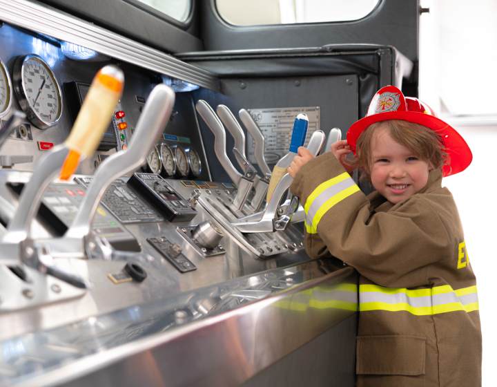This is a photo of a young boy wearing a fireman's jacket and a red firefighter helmet. He is smiling at the camera and playing with the levers inside the firetruck. 