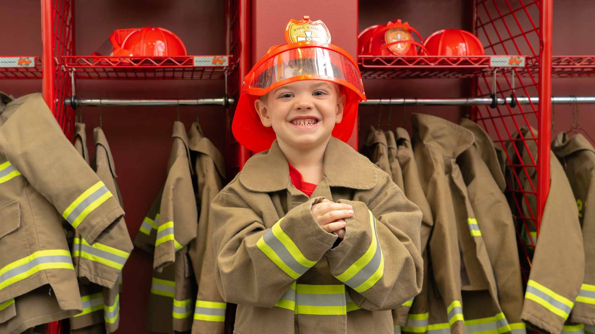 A photo of a boy smiling at the camera dressed in a fireman jacket and red firefighter helmet. 