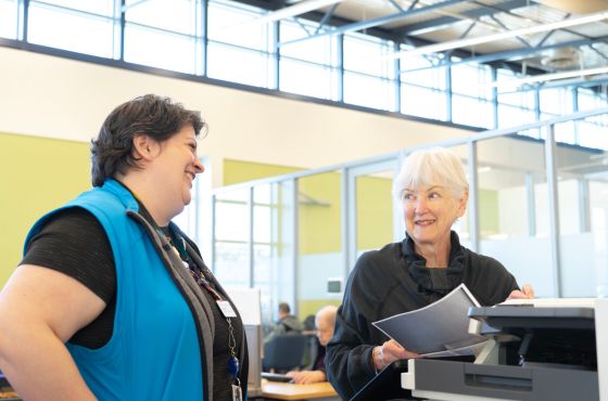 Two women in a library standing beside a printer. One is holding paper and one is wearing a blue vest.