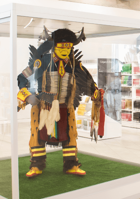 A traditional Indigenous regalia, featuring intricate beadwork and feathers, is displayed inside a glass case. The figure stands on a patch of artificial grass, creating a contrast between the natural elements of the costume and the modern display.