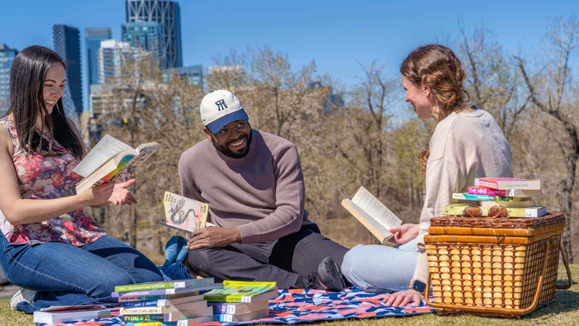A group of friends sitting on a picnic blanket with stacks of books all around them. They are all reading a book and smiling. 