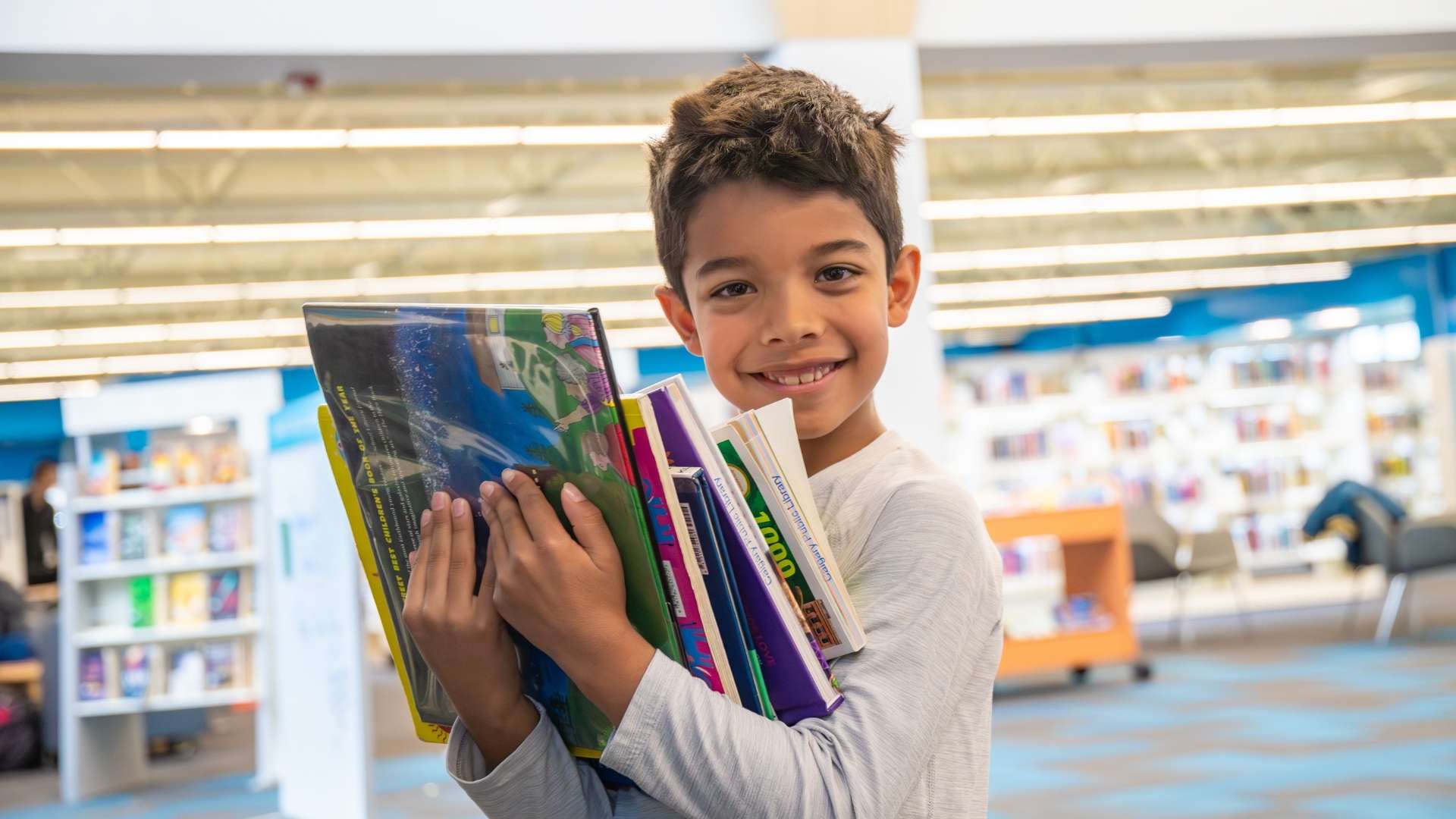 A child smiling at the camera holding a large stack of books. 