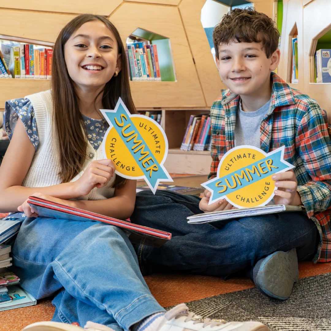 Two children sitting in a library reading nook hold Ultimate Summer Challenge signs, with books displayed around them.