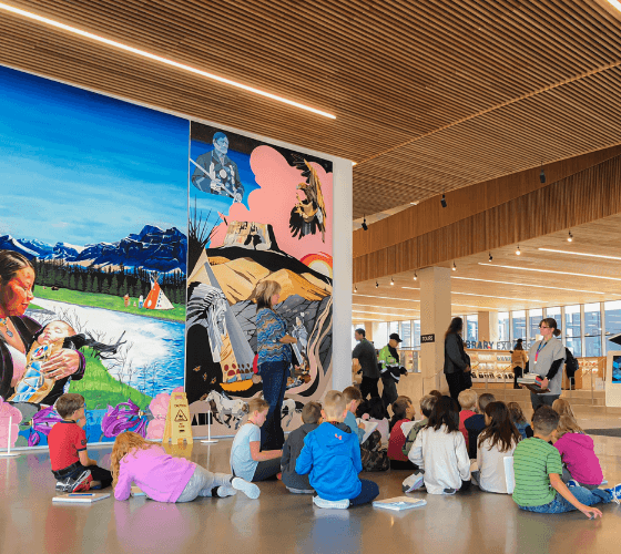 A series of painted panels depicts scenes of Indigenous life and nature. In front of it is school children, sitting on the floor and listening to a Library staff member.