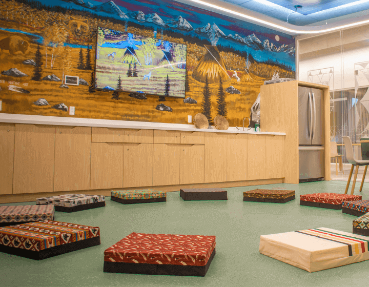 This is a photograph of the Indigenous Languages Resource Centre at Central Library. In the photograph, cushions are arranged in a circle on the floor. On the wall, there is a beautiful mural of a prairie landscape with mountains painted behind it.