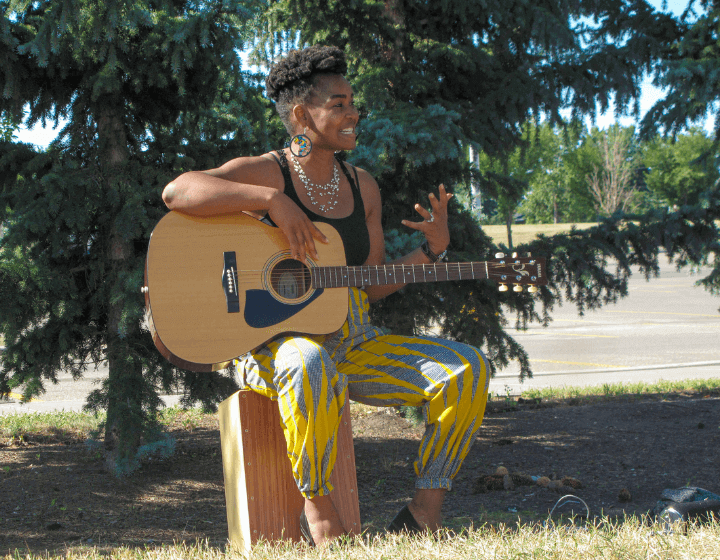 A photograph of an adult sitting on a wooden block in a park. They have an acoustic guitar on their lap and are smiling at the crowd.