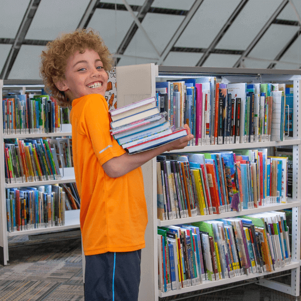 A child standing near a bookshelf holding a stack of books and smiling at the camera.